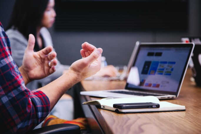 Person gesturing during a meeting with a laptop and notebook on the table.