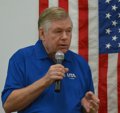 Man speaking into a microphone in front of an American flag.